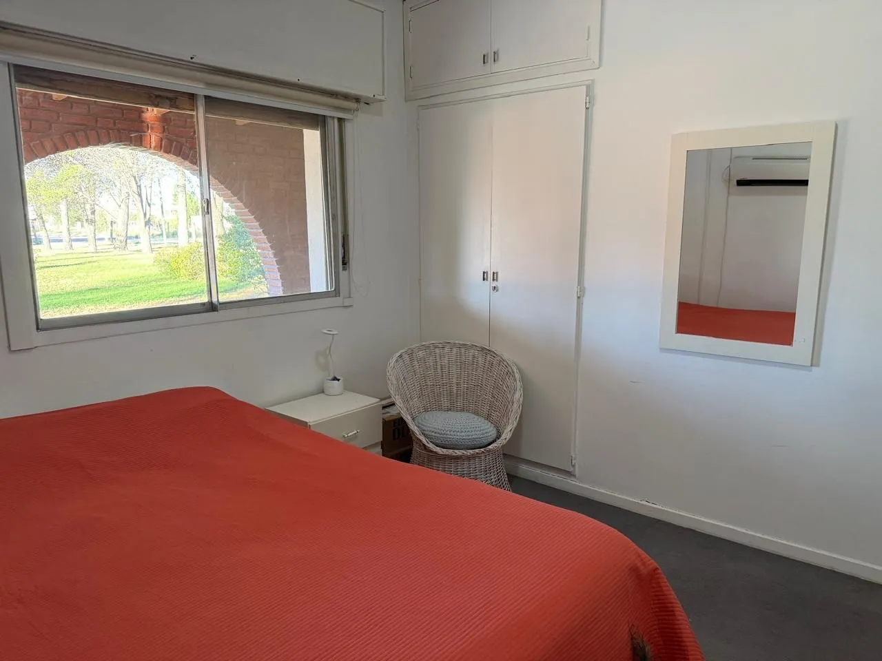 Dormitorio con cama de dos plazas, placard empotrado, un sillón de mimbre y una ventana que da a los arcos de la galería.